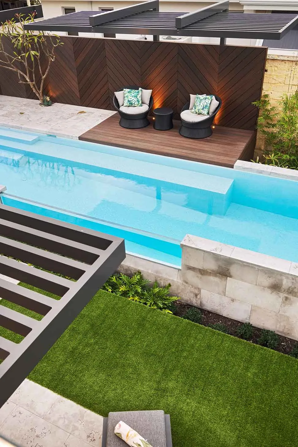 Elevated poolside view featuring a sleek blue swimming pool, green lawn, and a stylish wooden deck with cosy seating and warm lighting.