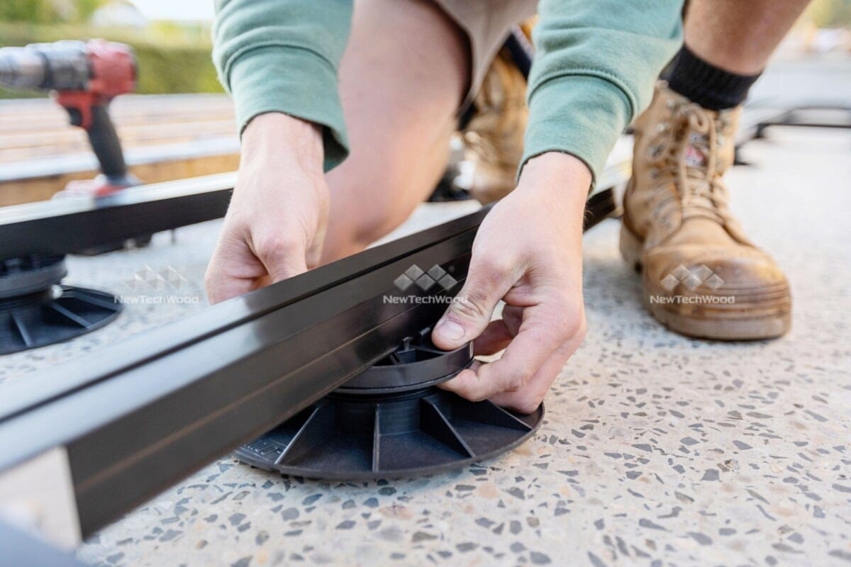 Close-up of a worker's hands adjusting a deck support pedestal while installing a NewTechWood deck on a terrazzo surface, with tools visible in the background.