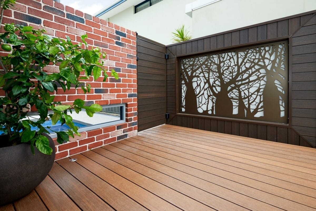 Modern outdoor patio with composite deck, a decorative metal privacy screen featuring tree silhouettes, and a red brick wall with a built-in rectangular window.
