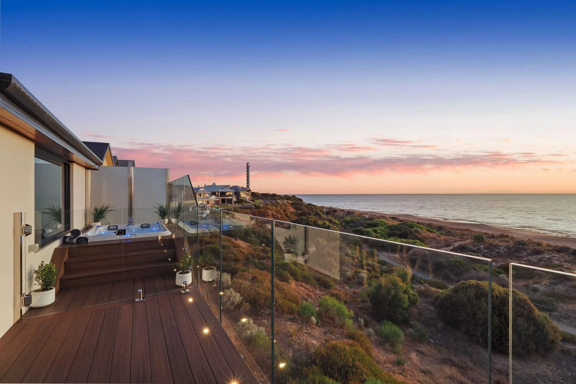 Luxurious seaside composite decking with integrated lighting on a modern home's terrace, overlooking a tranquil beach and lighthouse during sunset.