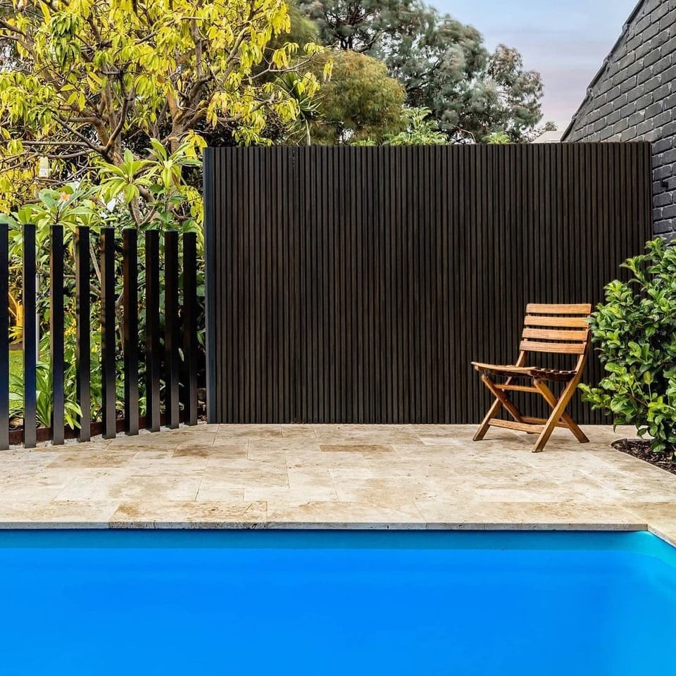 Outdoor pool area with a blue-tiled pool, a wooden folding chair, and a dark vertical slat fence surrounded by lush greenery.
