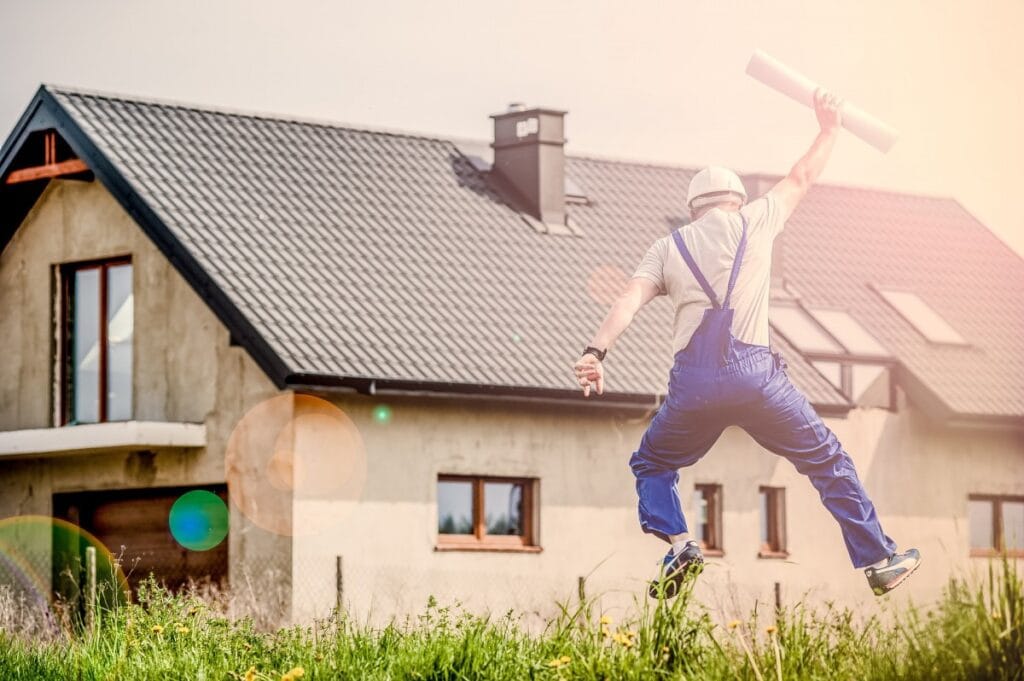 Construction worker in blue overalls jumping in excitement while holding building plans in front of a newly constructed house