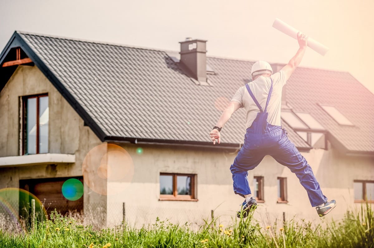 Construction worker in blue overalls jumping in excitement while holding building plans in front of a newly constructed house