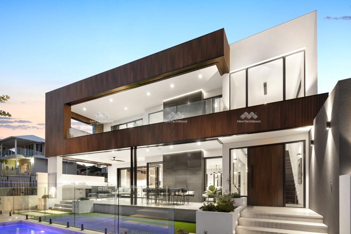 Modern luxury home with large glass windows, wood panelling, and a spacious balcony overlooking a pool at sunset