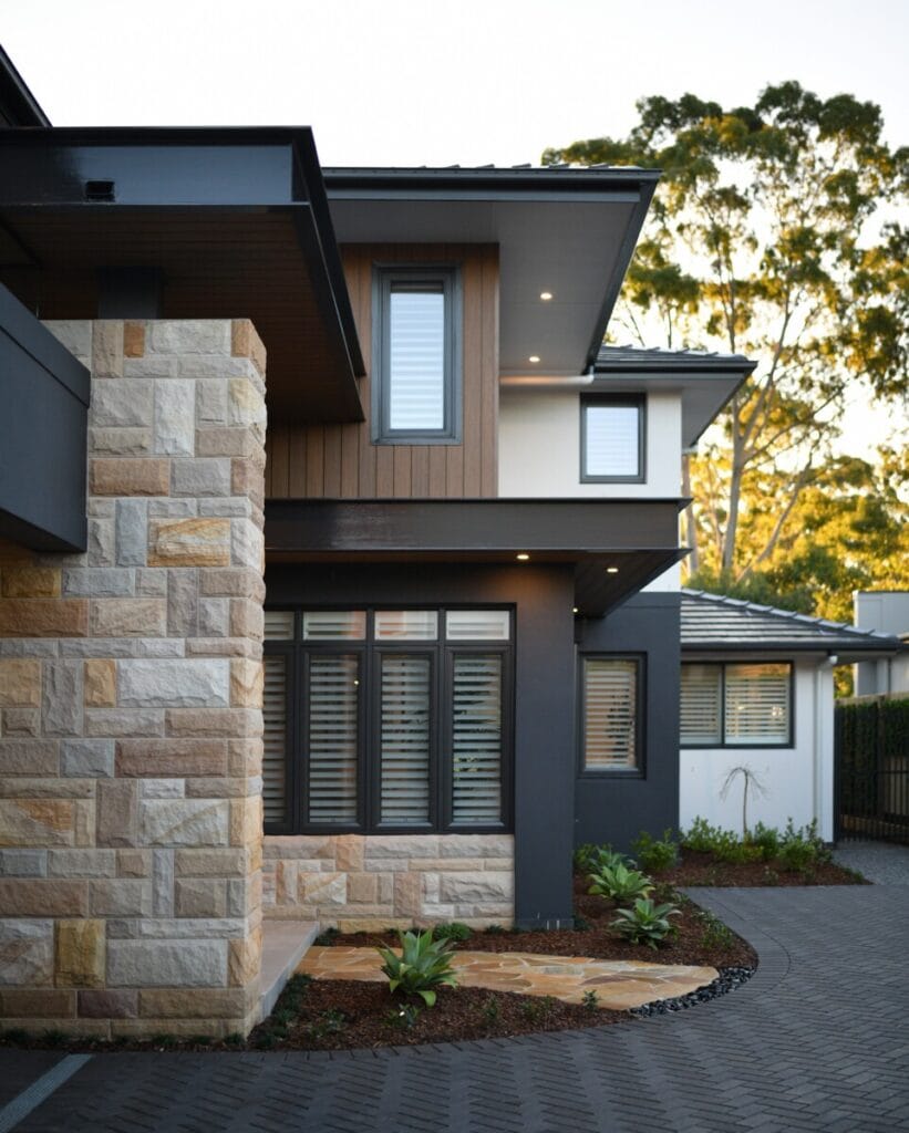 Modern home exterior with stone and wood cladding, featuring large windows with shutters and a landscaped garden.