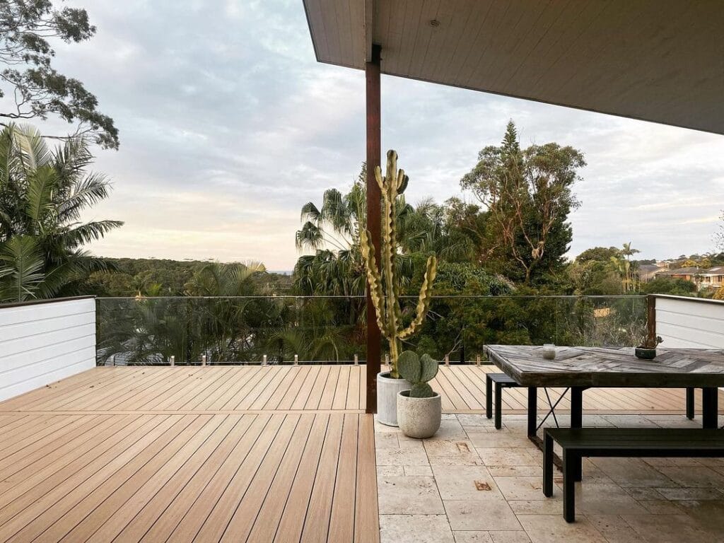 Spacious outdoor area with NewTechWood decking, featuring a modern black table and benches, potted cacti, and a view of lush greenery under a cloudy sky.