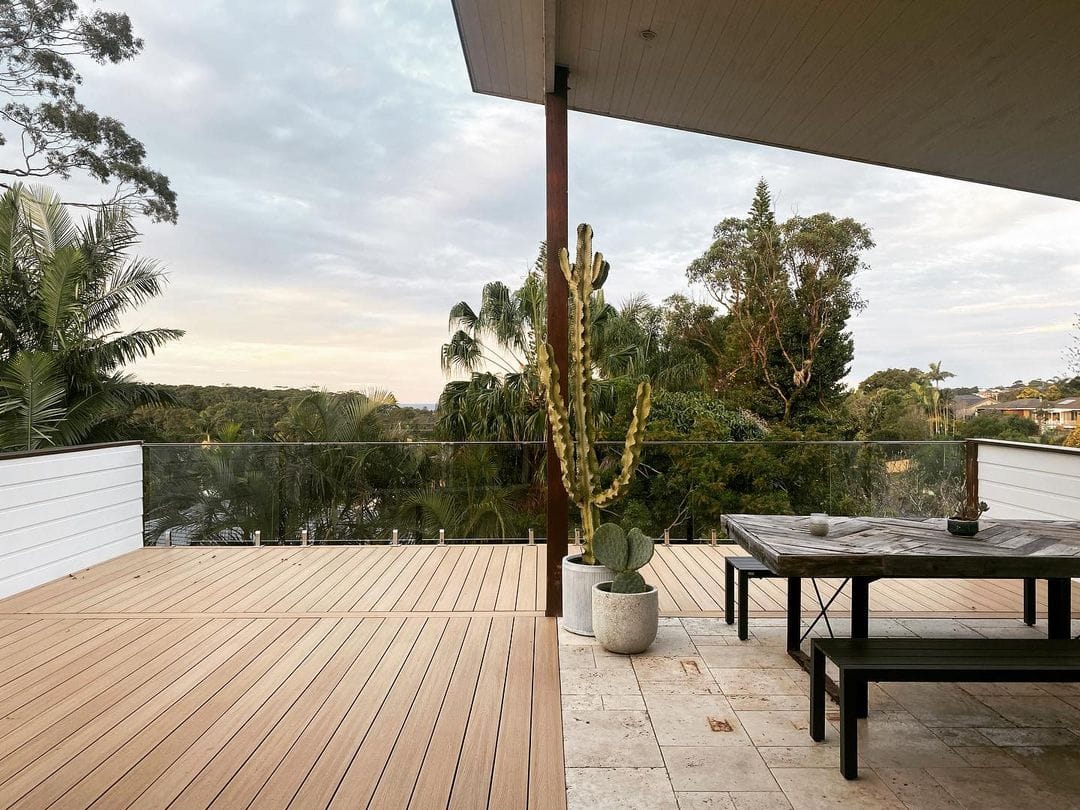 Spacious outdoor area with NewTechWood decking, featuring a modern black table and benches, potted cacti, and a view of lush greenery under a cloudy sky.