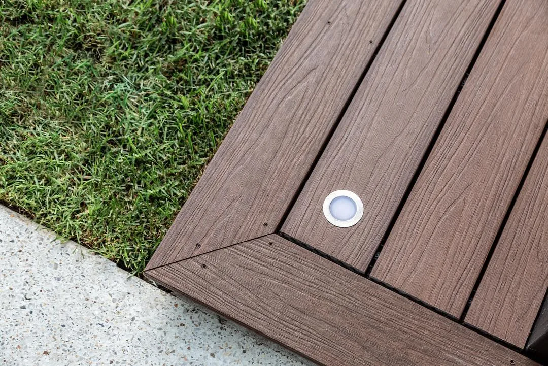 Close-up of a small round deck light installed in a brown composite deck board, providing subtle illumination and enhancing outdoor aesthetics.