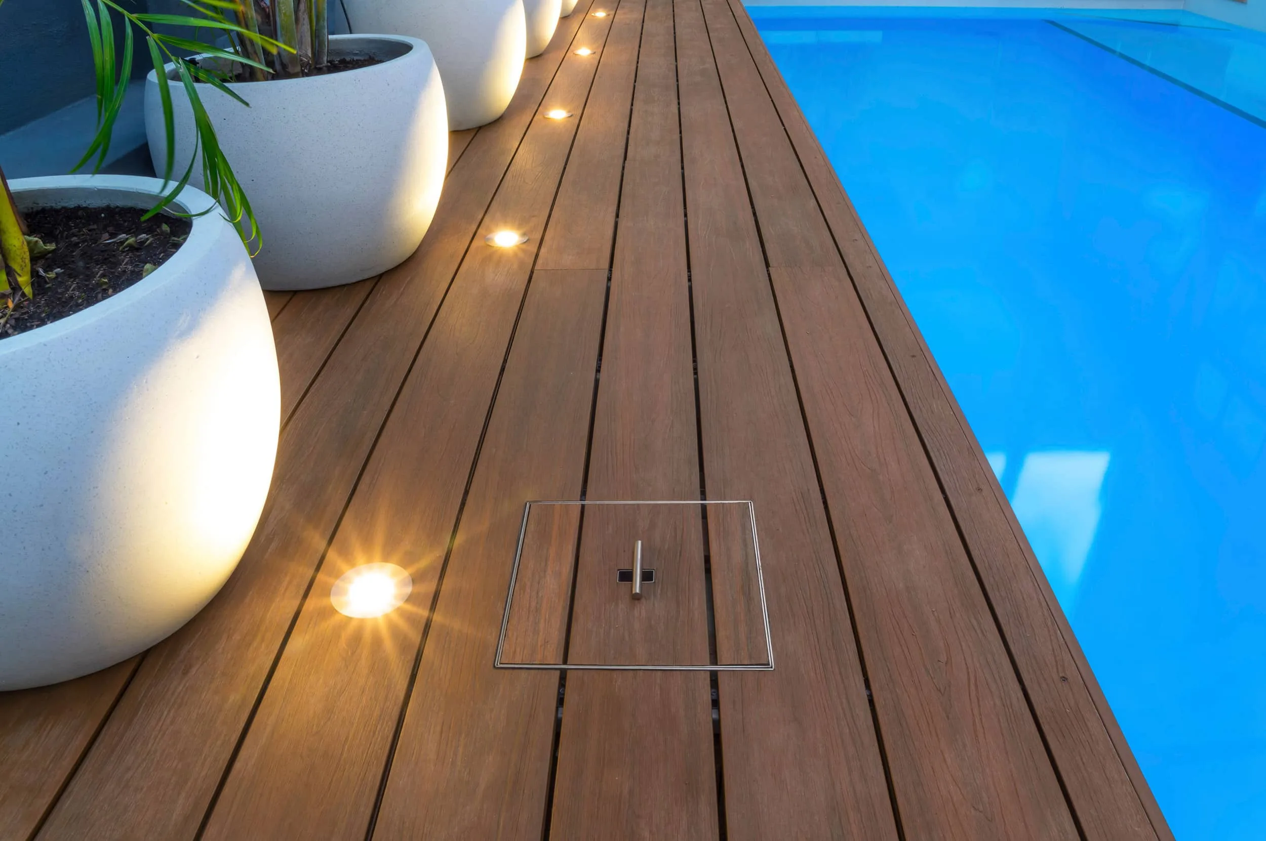 A sleek wooden poolside deck with integrated lighting, large planters, and a concealed access panel.