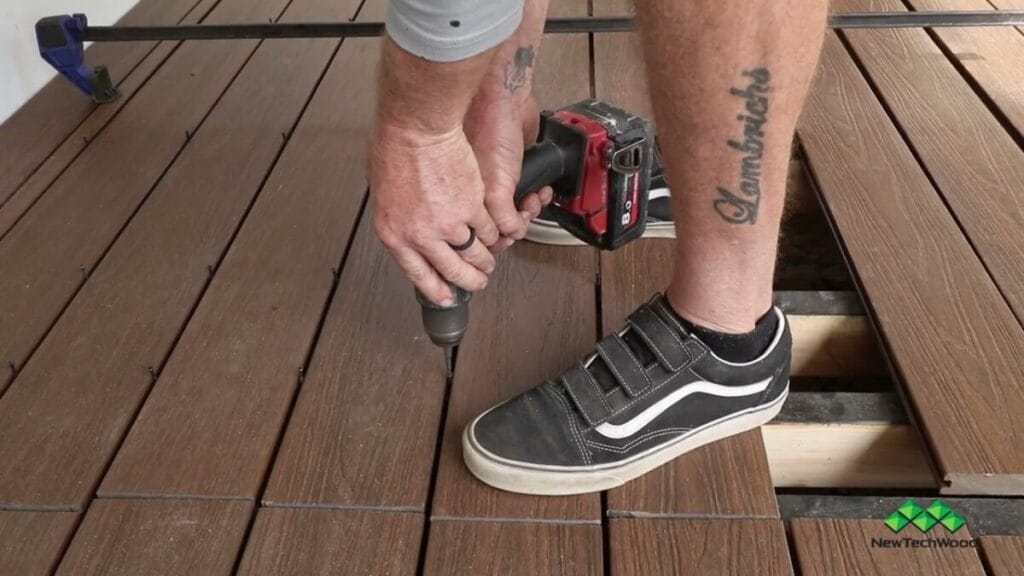 Close-up of a person installing composite decking using a power drill, with visible tattooed arm and foot in a black sneaker