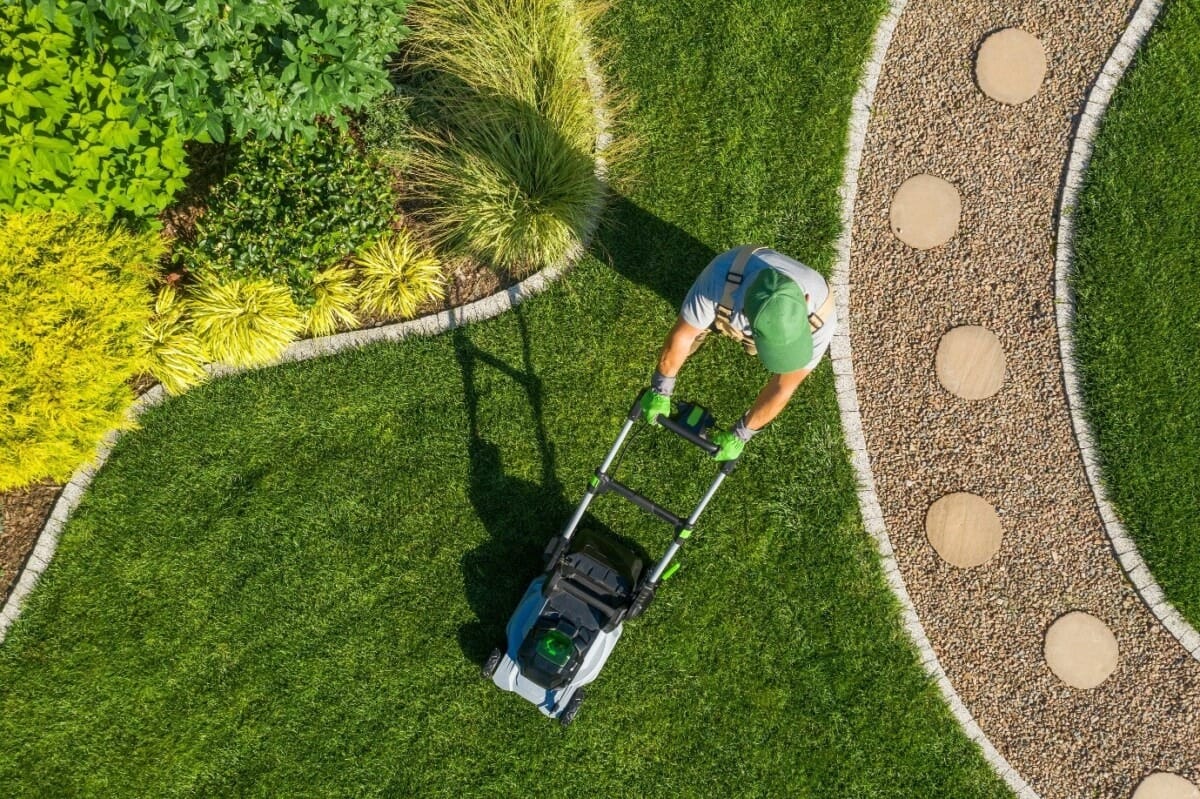 Aerial view of a gardener mowing a well-maintained lawn with a modern lawn mower, surrounded by landscaped garden beds and a stone pathway