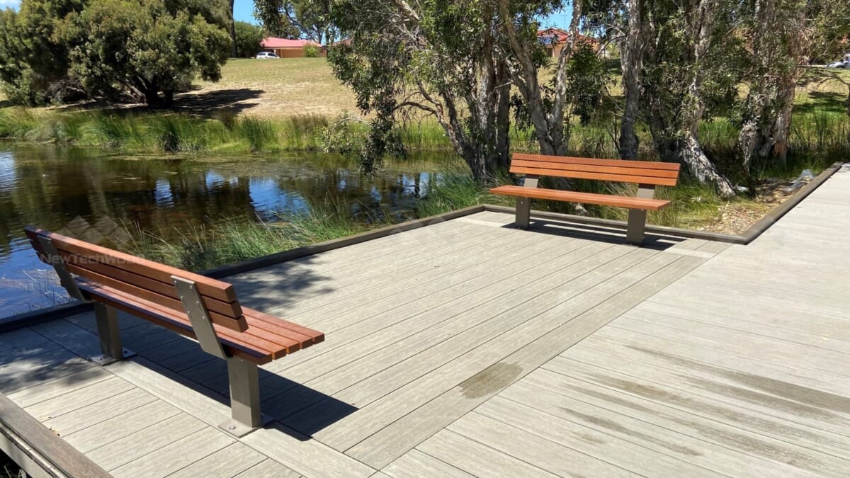 Two wooden benches in the City of Mandurah, set on NewTechWood composite decking and surrounded by a peaceful pond bordered by trees and grassy terrain.