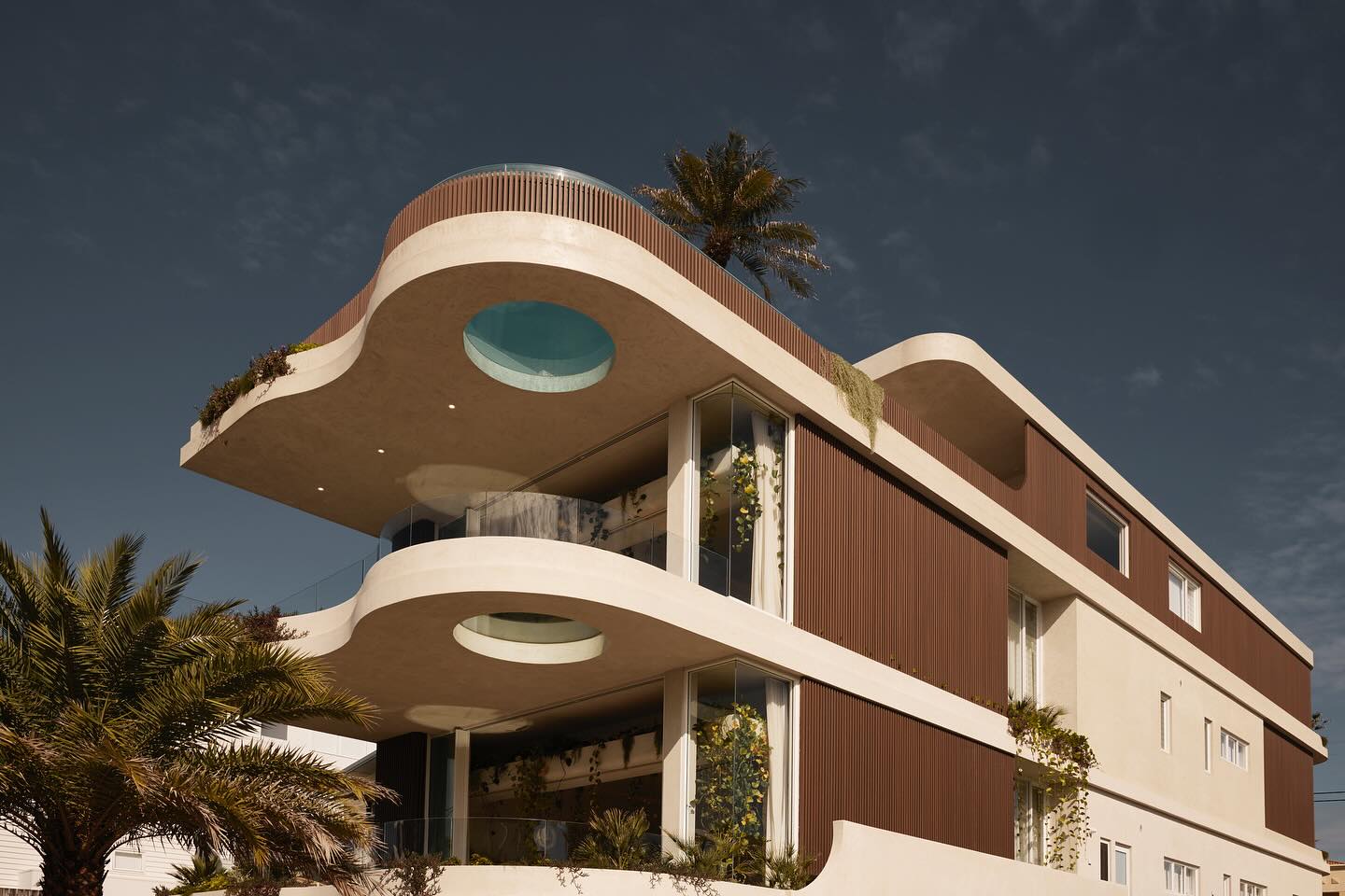 This multi-story luxury home boasts a distinctive sculptural design, with sweeping organic lines, vertical timber cladding, and strategically placed greenery. The signature feature is a glass-bottomed circular cutout, allowing natural light to filter through different levels. This breathtaking residence redefines modern coastal architecture.