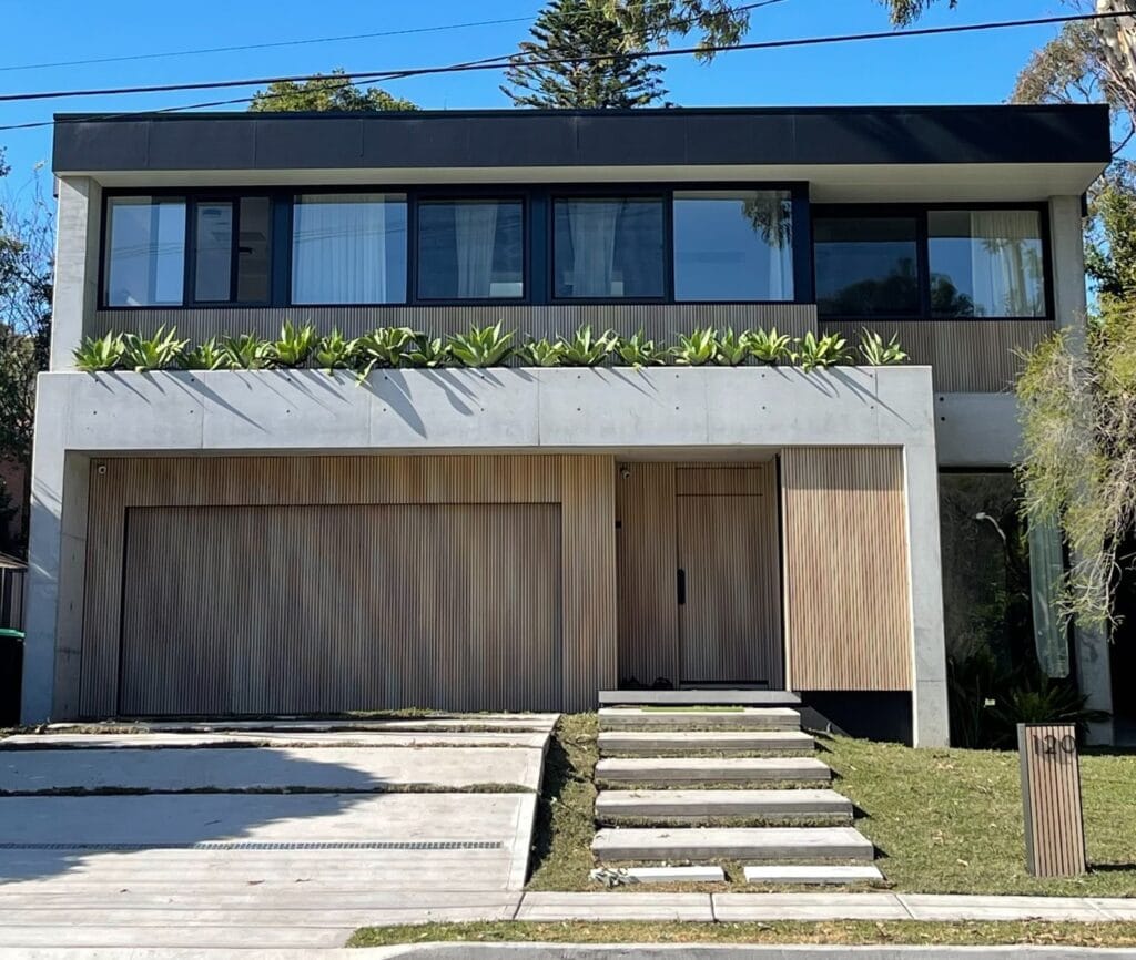 A contemporary two-story home featuring wood cladding, concrete finishes, black window frames, and lush greenery on the facade.